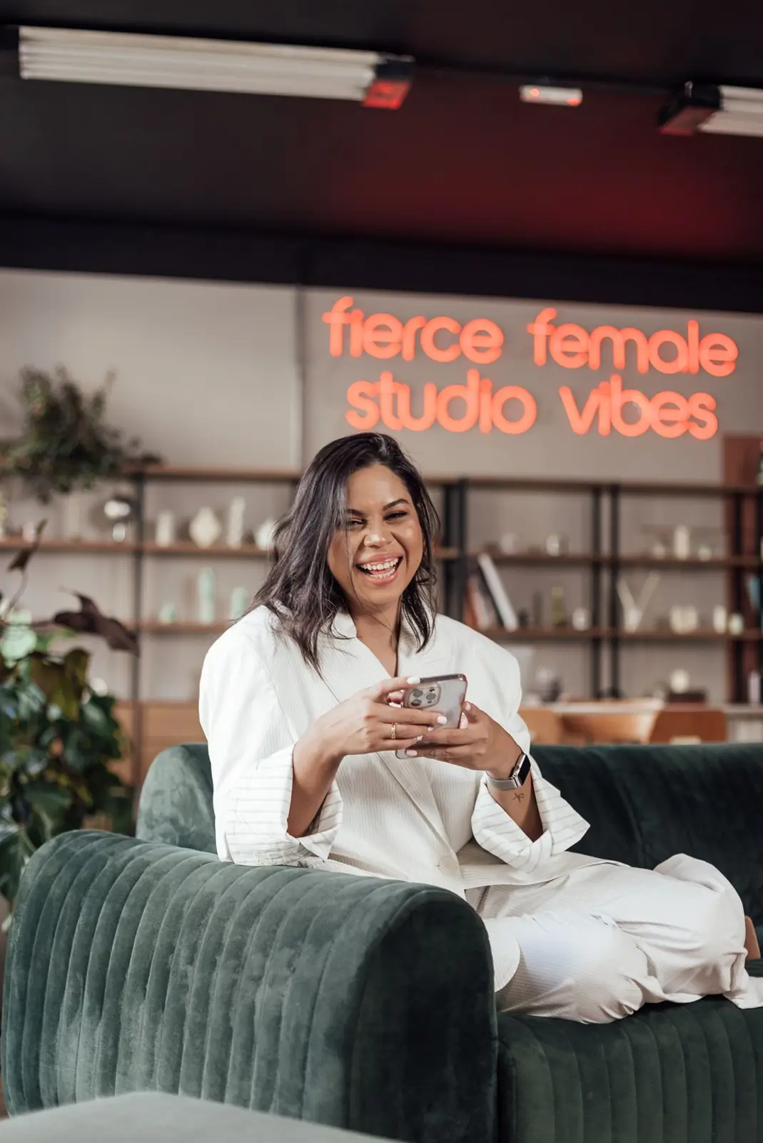 Bruna dos Santos de terno branco, sentada em um sofá verde-escuro, sorrindo