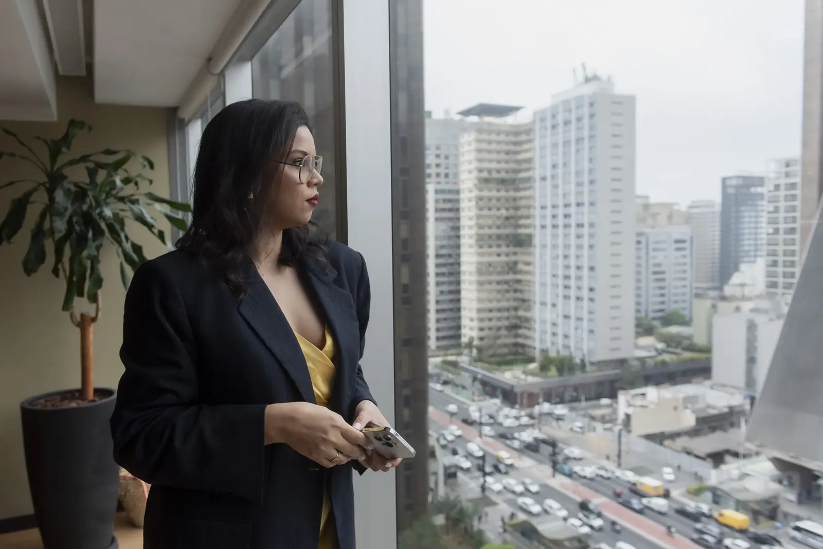 Bruna dos Santos de blazer preto, diante de uma janela com vista para a cidade