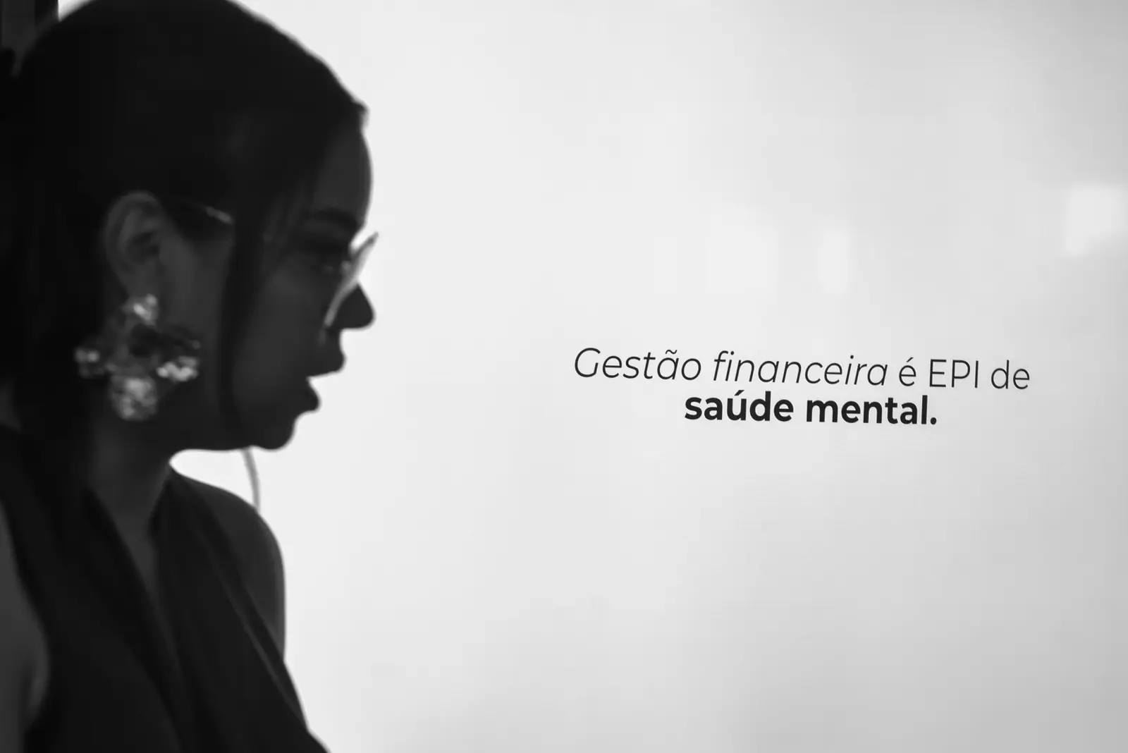 Bruna de perfil diante de um telão com a frase: Gestão financeira é EPI de saúde mental.